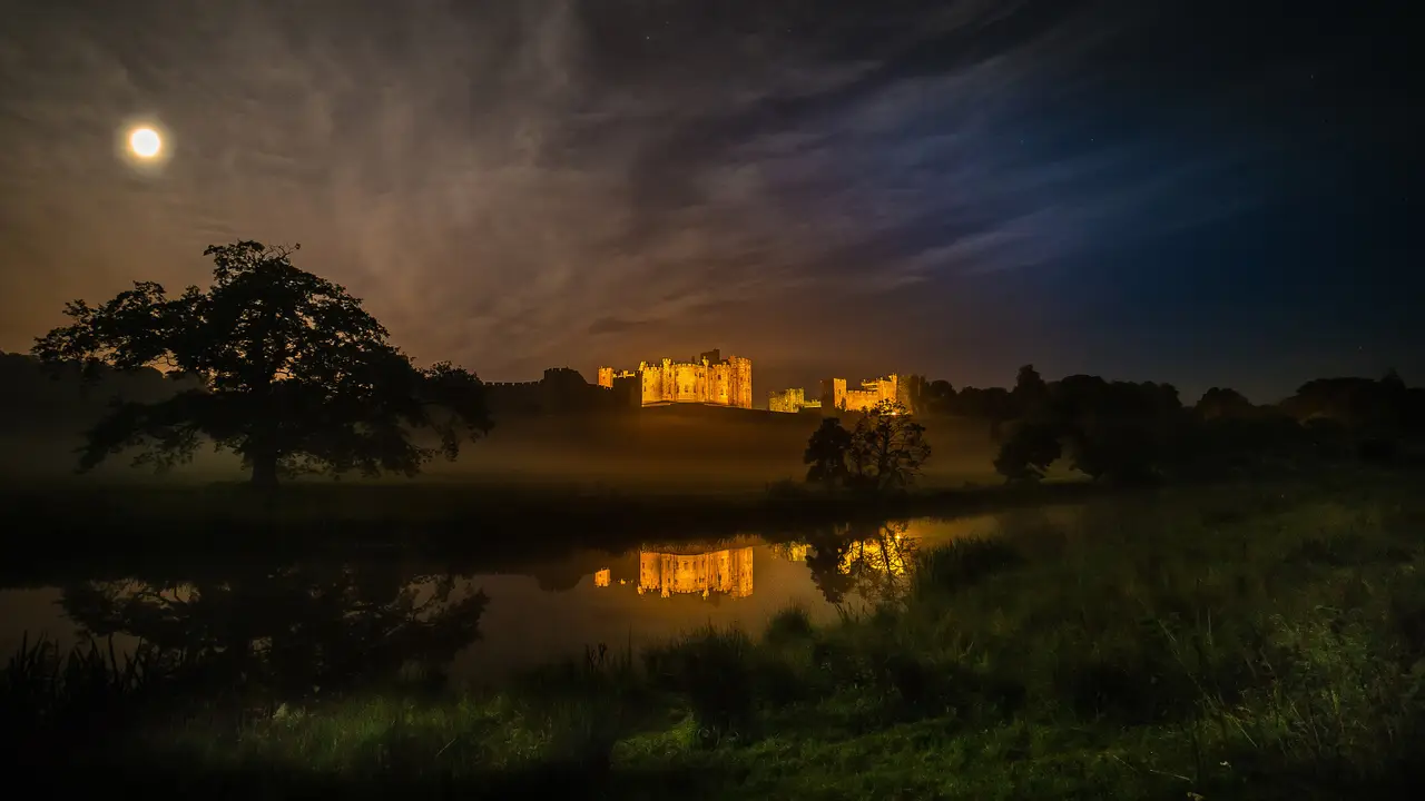 Alnwick Castle, a Hogwarts filming location, lit up under a full moon with mist over the river
