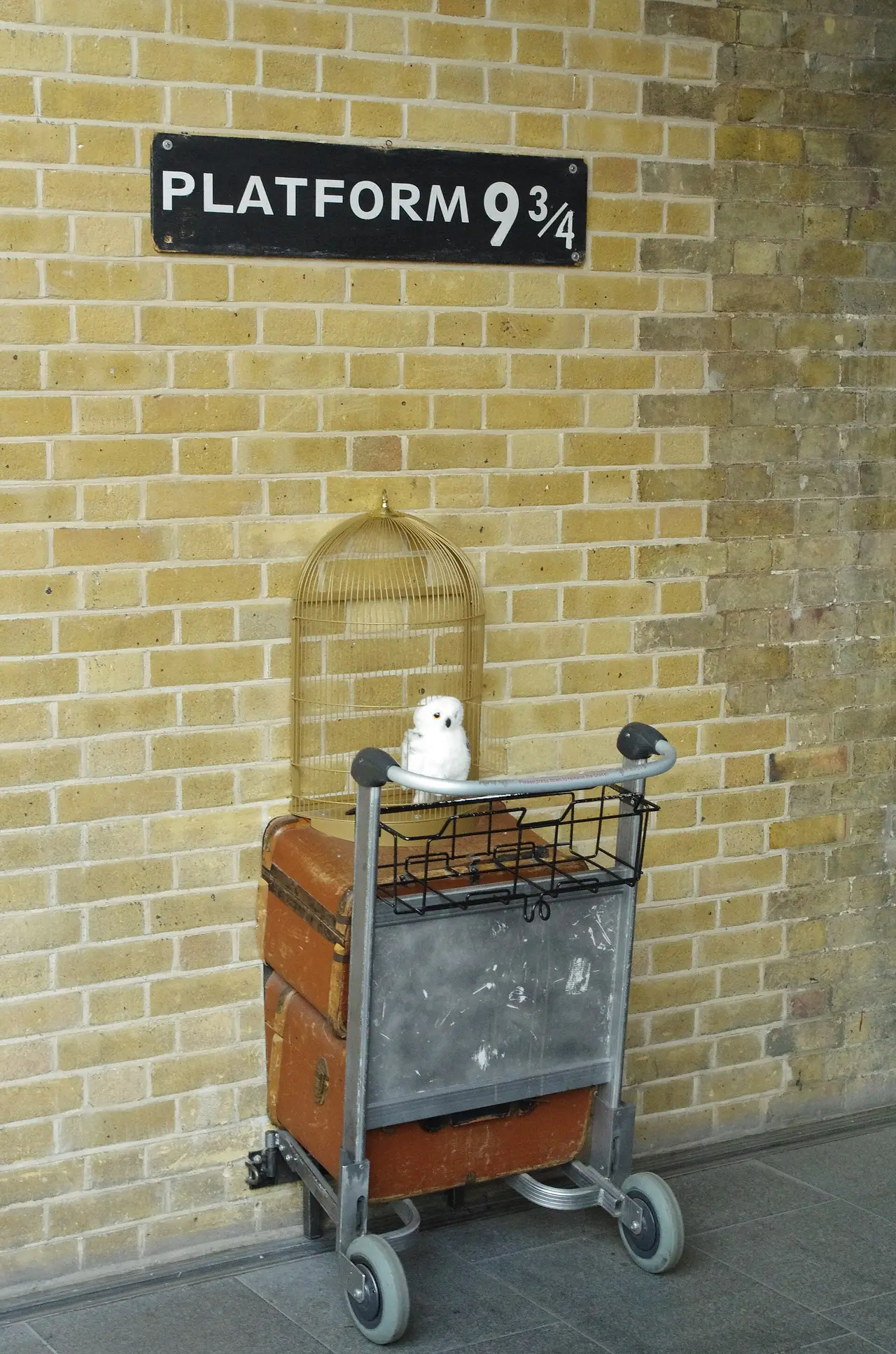 A Platform 9¾ sign above a luggage trolley and owl cage on a brick wall